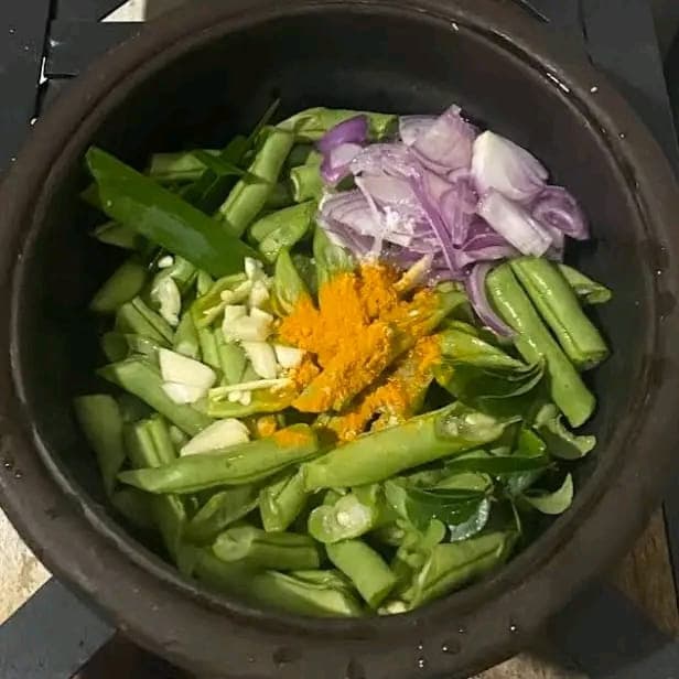A clay pot of green beans, red onion, garlic, pandan leaf and curry leaves before cooking